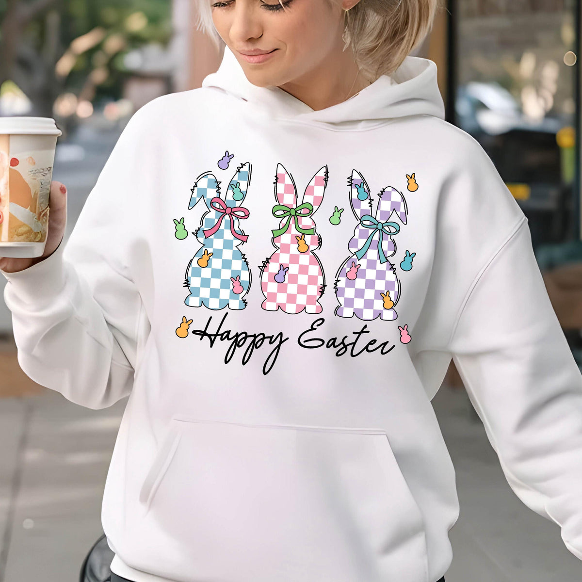 May include: White hoodie with a graphic of three pastel colored bunnies with checkered patterns and bows. The bunnies are surrounded by small pastel colored eggs. The text "Happy Easter" is printed below the bunnies.