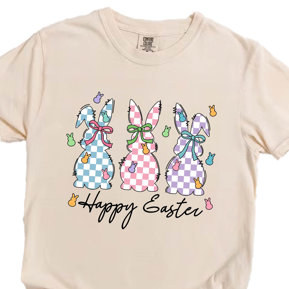 May include: A beige t-shirt with a graphic of three pastel checkered bunnies and the text "Happy Easter".