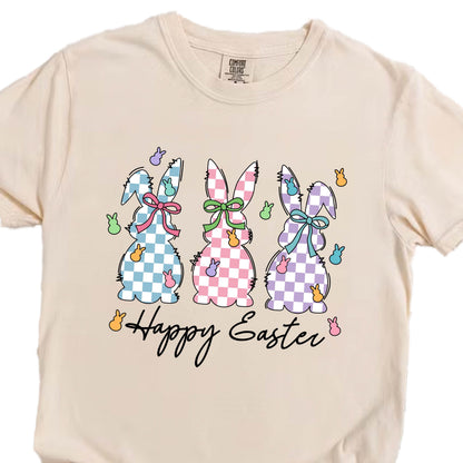May include: A beige t-shirt with a graphic of three pastel checkered bunnies and the text "Happy Easter".