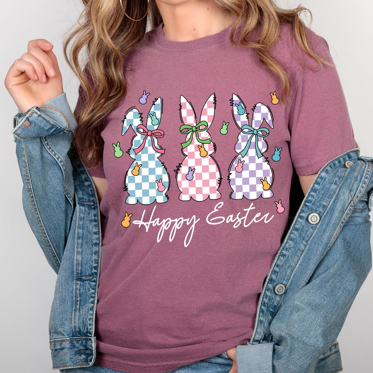 May include: A mauve t-shirt with a graphic of three Easter bunnies in a row. The bunnies are checkered in blue, pink, and purple. Each bunny has a bow and is surrounded by small yellow chicks. The text "Happy Easter" is below the bunnies.