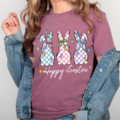 May include: A mauve t-shirt with a graphic of three Easter bunnies in a row. The bunnies are checkered in blue, pink, and purple. Each bunny has a bow and is surrounded by small yellow chicks. The text "Happy Easter" is below the bunnies.