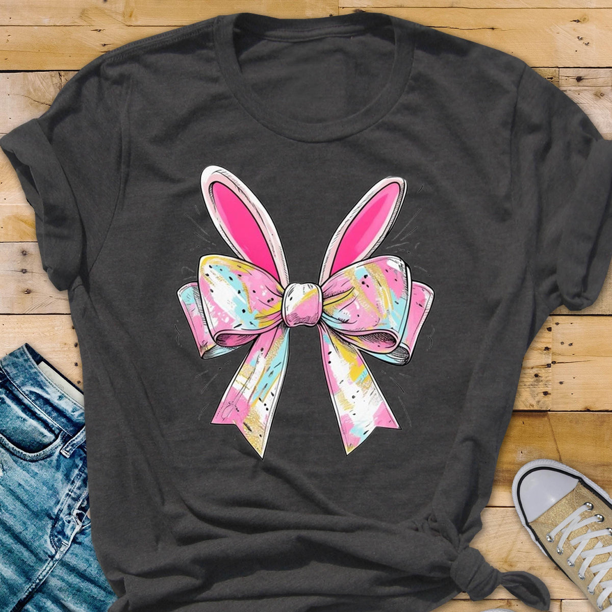 May include: A dark gray t-shirt with a pink and white bow with bunny ears. The bow has a watercolor-like design with pink, blue, and yellow colors.