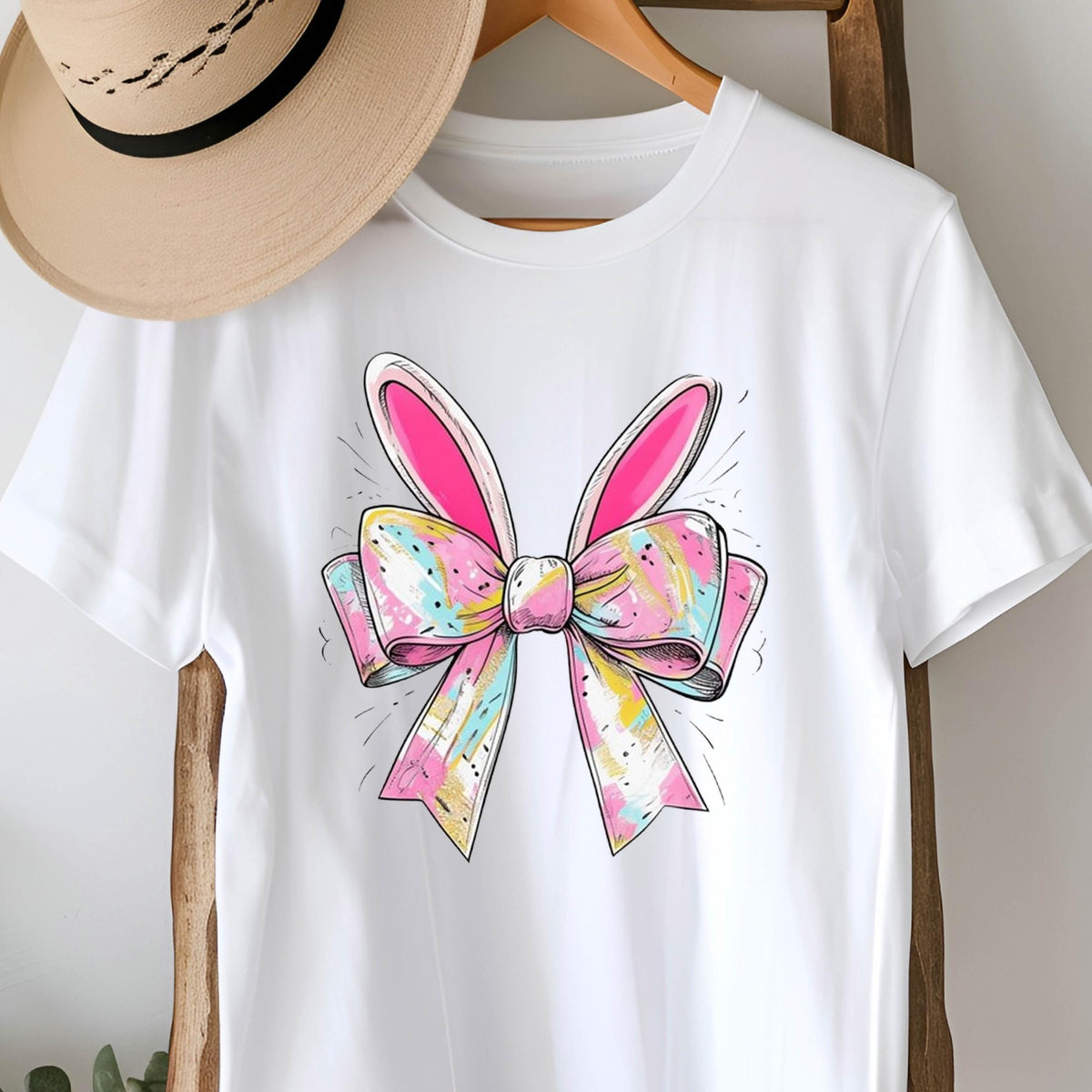 May include: White t-shirt with a colorful bow with bunny ears. The bow is pink, blue, yellow, and white.