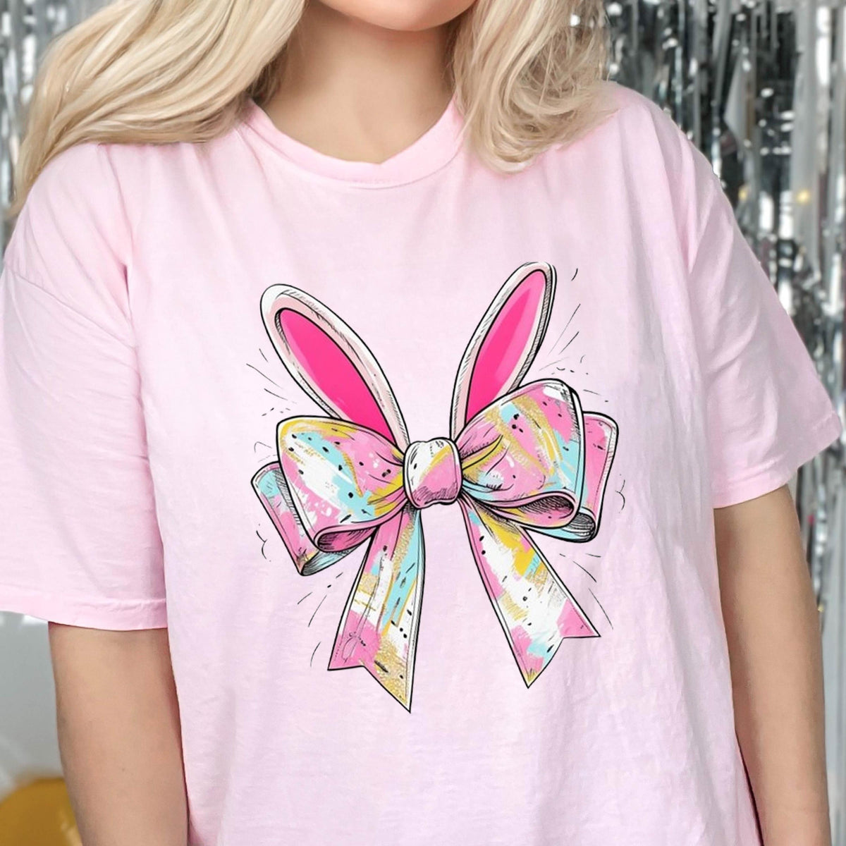 May include: Pink t-shirt with a colorful bow and bunny ears design. The bow is tied with a yellow ribbon and has pink, blue, and yellow stripes.