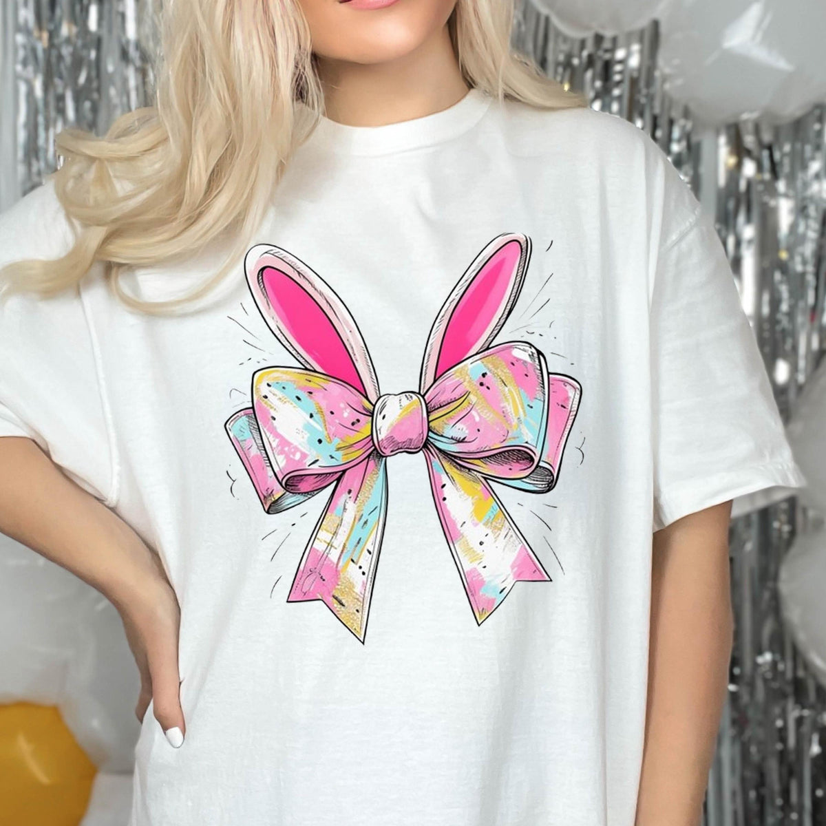 May include: White t-shirt with a pink and white bow with bunny ears. The bow has a colorful, abstract pattern.