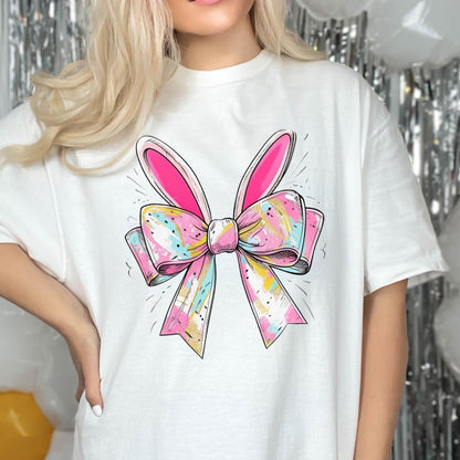 May include: White t-shirt with a pink and white bow with bunny ears. The bow has a colorful, abstract pattern.