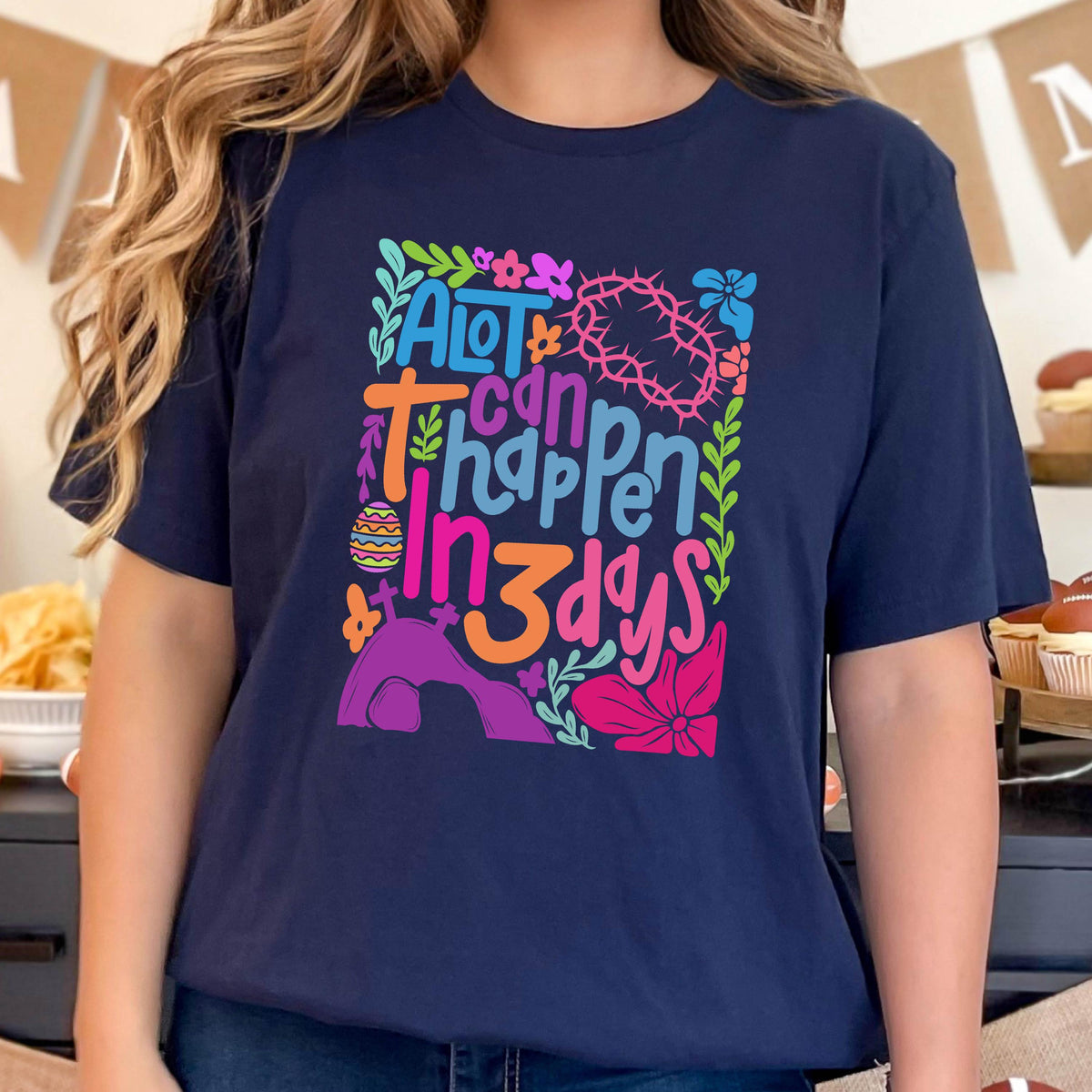 May include: A navy blue t-shirt with a colorful graphic design featuring the text "A lot can happen in 3 days". The graphic includes flowers, a crown of thorns, and an Easter egg.