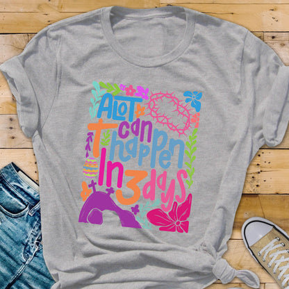 May include: A gray t-shirt with a colorful graphic design that says "A lot can happen in 3 days". The graphic includes flowers, a cross, a crown of thorns, and a tomb.