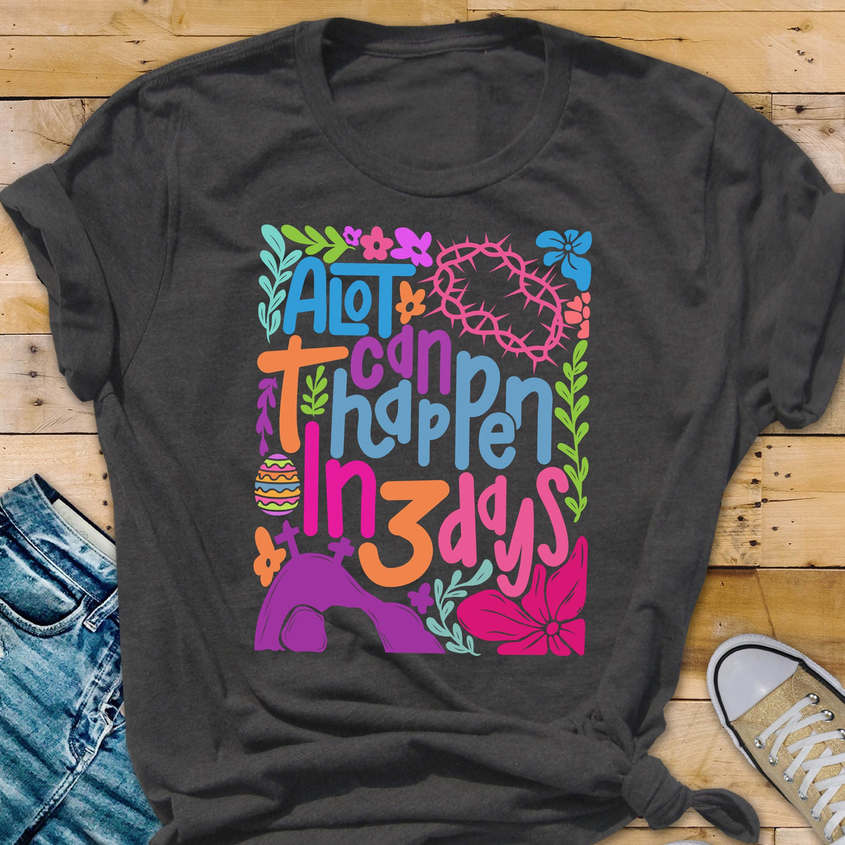 May include: A dark gray t-shirt with a colorful graphic design that says "A lot can happen in 3 days". The graphic includes flowers, a crown of thorns, and a tomb.