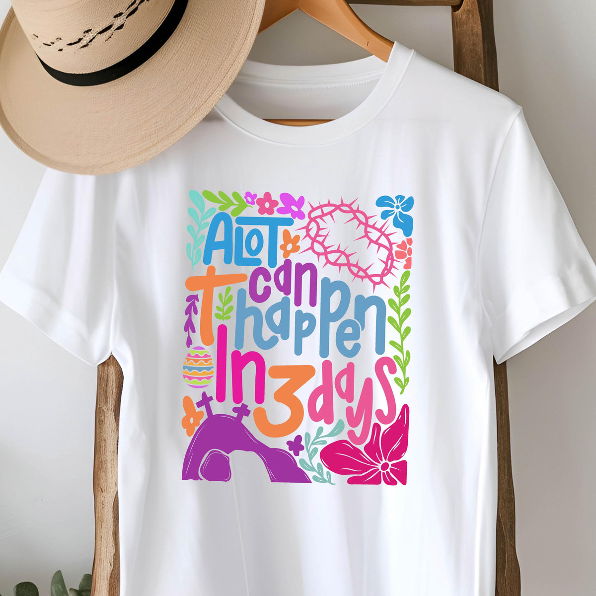 May include: White t-shirt with a colorful graphic design that says "A lot can happen in 3 days". The design includes flowers, a crown of thorns, a cross, and an Easter egg.