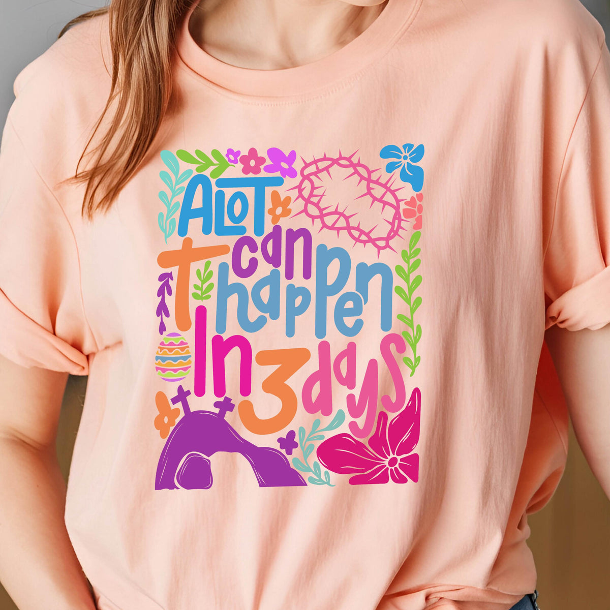 May include: A pink t-shirt with a colorful graphic design that says "A lot can happen in 3 days". The graphic includes flowers, a crown of thorns, and a tomb.
