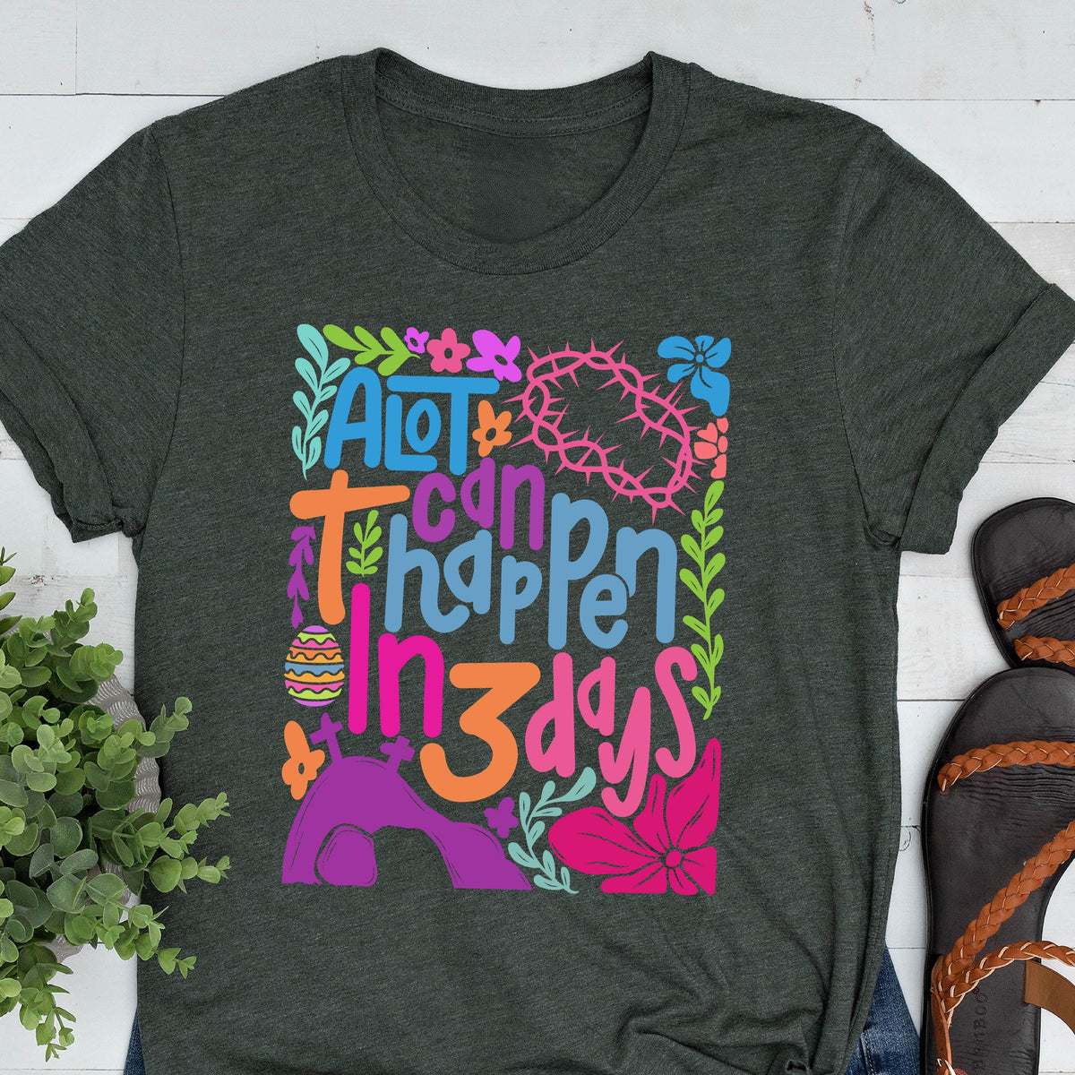 May include: A dark green t-shirt with a colorful graphic design that says "A lot can happen in 3 days". The graphic includes flowers, a crown of thorns, and an Easter egg.
