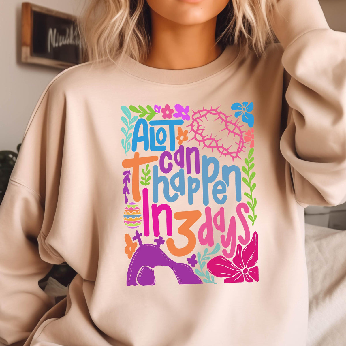 May include: A light beige sweatshirt with a colorful graphic design that says "A lot can happen in 3 days". The graphic includes flowers, an Easter egg, a cross, and a tomb.