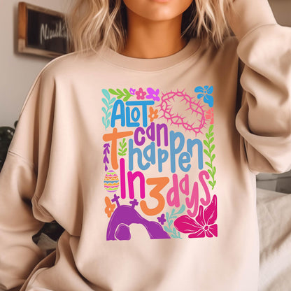 May include: A light beige sweatshirt with a colorful graphic design that says "A lot can happen in 3 days". The graphic includes flowers, an Easter egg, a cross, and a tomb.