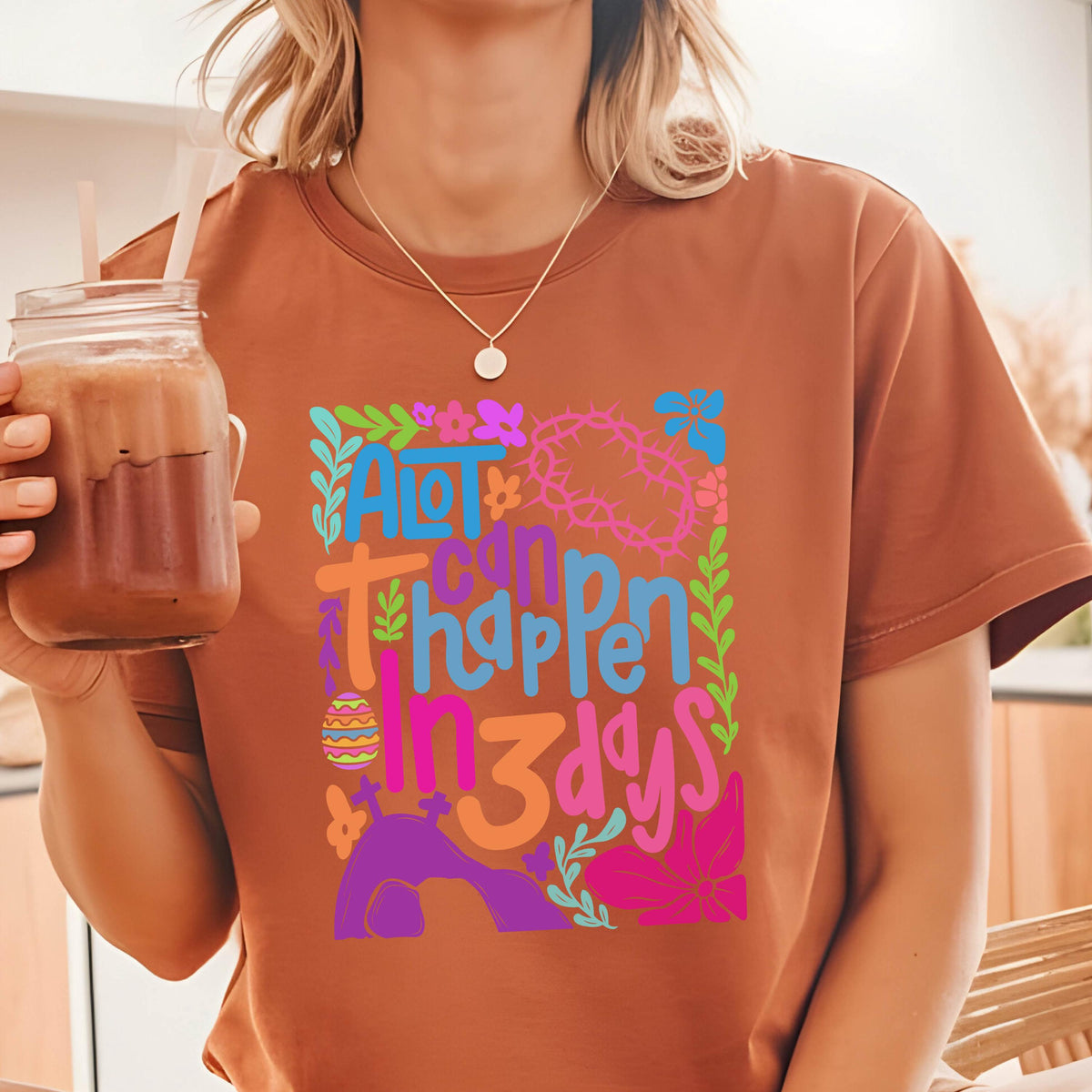 May include: A brown t-shirt with a colorful graphic design that says "A lot can happen in 3 days". The graphic includes a cross, a crown of thorns, an Easter egg, and a tomb.