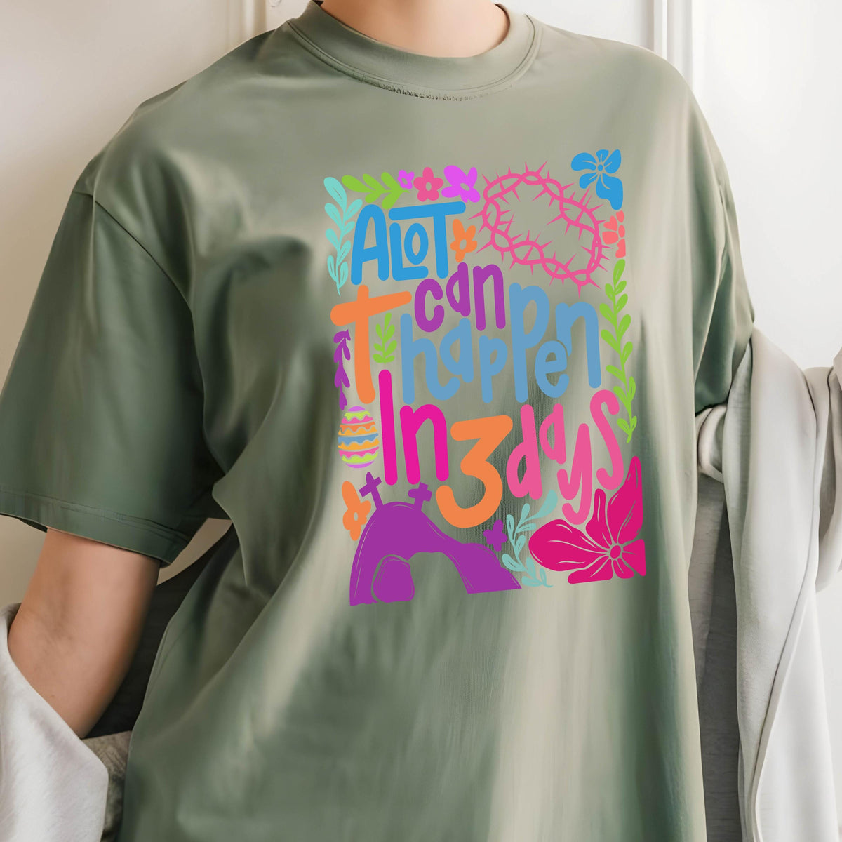 May include: A green t-shirt with a colorful graphic design that says "A lot can happen in 3 days". The design includes flowers, a cross, a crown of thorns, and an Easter egg.