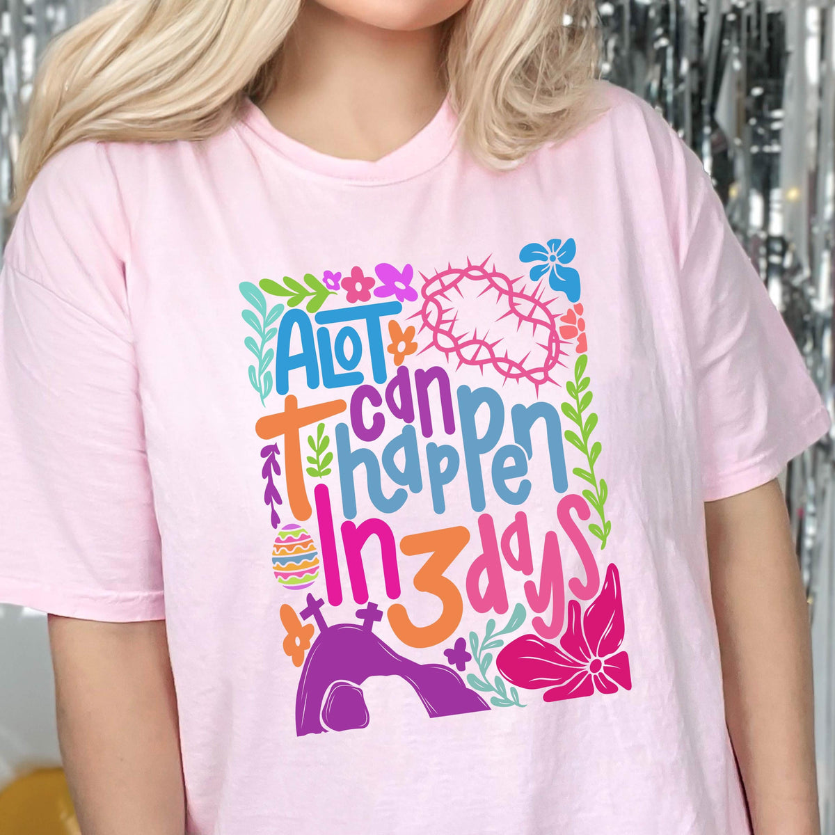 May include: Pink t-shirt with a colorful graphic design that says "A lot can happen in 3 days". The design features flowers, a crown of thorns, and a tomb.
