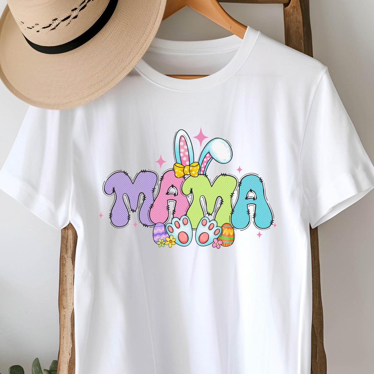 May include: White t-shirt with a colorful graphic of the word "MAMA" in a playful font. The graphic includes bunny ears, a bow, and Easter eggs.