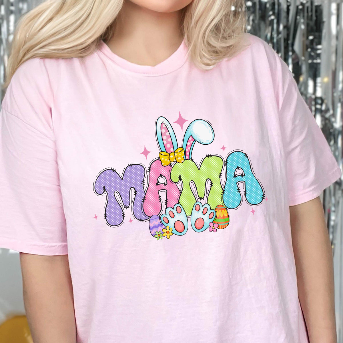 May include: A light pink t-shirt featuring the word "MAMA" in a colorful, bubble-style font. The design includes bunny ears, Easter eggs, and paw prints, creating a festive Easter theme.