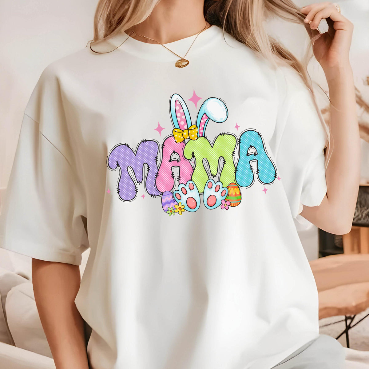 May include: White t-shirt with a colorful "MAMA" graphic. The design features bunny ears, Easter eggs, and flower accents. The letters are in purple, green, and blue with a playful font, perfect for Easter.
