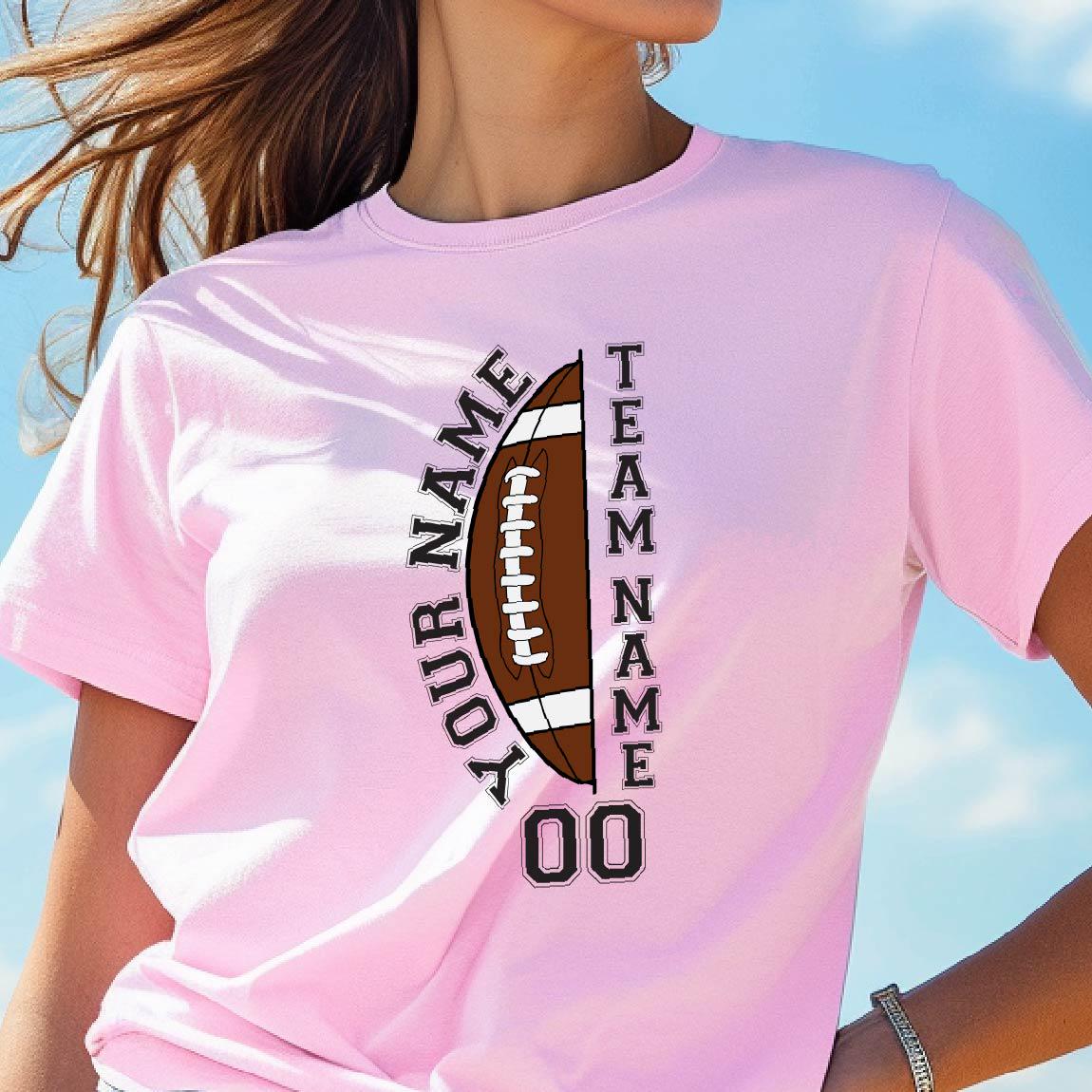May include: A light pink t-shirt featuring a graphic of a brown football split in half. The text YOUR NAME is printed vertically on the left, TEAM NAME on the right, and the number 00 is printed in black at the bottom of the football graphic.