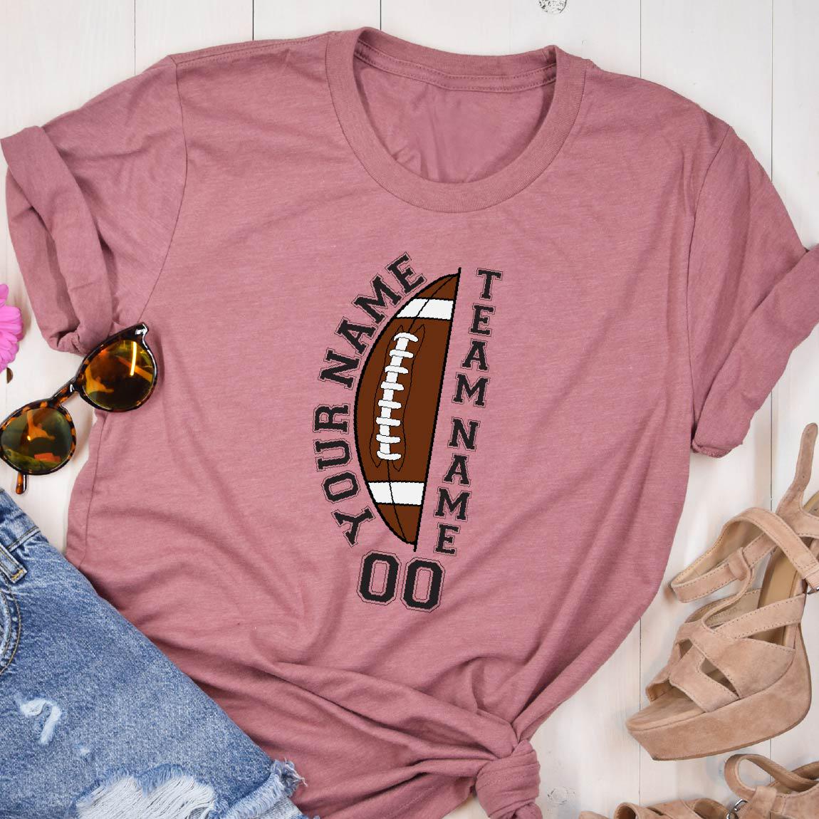 May include: A mauve short-sleeved t-shirt featuring a graphic of a brown American football with white laces. The text "YOUR NAME" and "TEAM NAME" is printed vertically alongside the football, with the number "00" displayed at the bottom.