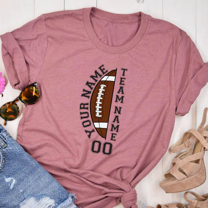 May include: A mauve short-sleeved t-shirt featuring a graphic of a brown American football with white laces. The text "YOUR NAME" and "TEAM NAME" is printed vertically alongside the football, with the number "00" displayed at the bottom.