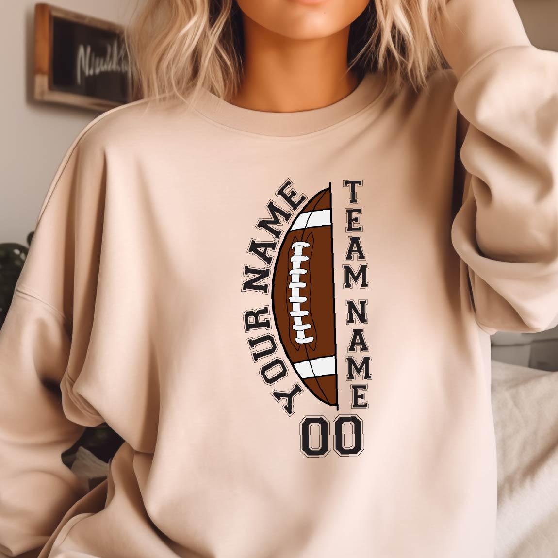 May include: A beige sweatshirt featuring a brown and white football graphic. The football design includes the text "YOUR NAME" and "TEAM NAME" vertically, with the number "00" at the bottom. The sweatshirt is a neutral color.