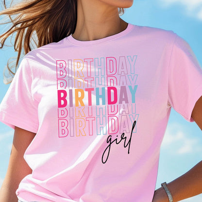 May include: A light pink t-shirt with the word "BIRTHDAY" stacked in a colorful, repeating pattern. The word "girl" is written in a cursive font below the stacked text. The shirt is short-sleeved.
