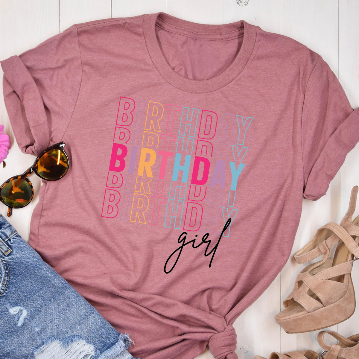 May include: Dusty rose-colored t-shirt with the word "BIRTHDAY" in colorful vertical text and the word "girl" in a cursive font. The shirt is folded, with sunglasses and denim shorts visible.