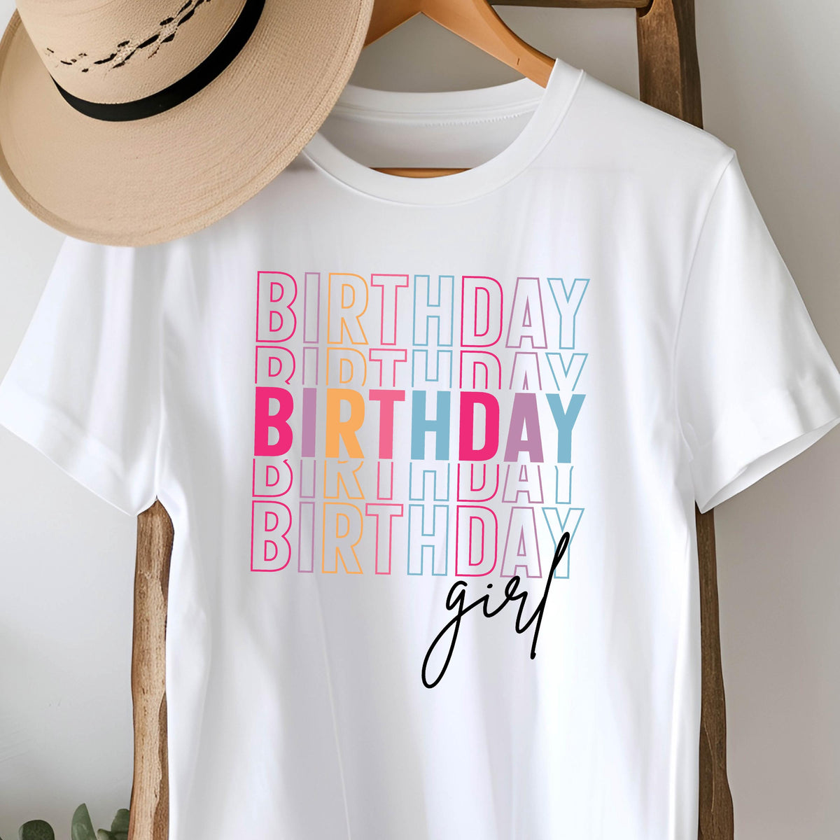 May include: White t-shirt with the word "BIRTHDAY" stacked in colorful text, with the word "girl" in cursive script below. The text is in pink, orange, blue, and teal. A tan hat hangs on a wooden hanger.