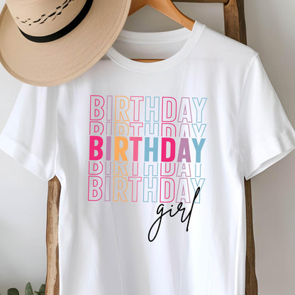 May include: White t-shirt with the word "BIRTHDAY" stacked in colorful text, with the word "girl" in cursive script below. The text is in pink, orange, blue, and teal. A tan hat hangs on a wooden hanger.
