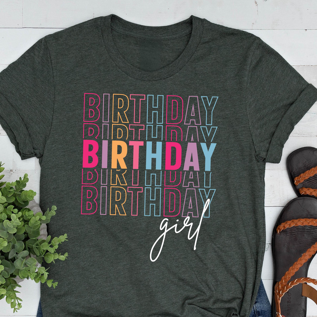 May include: Dark gray t-shirt with the word "BIRTHDAY" stacked multiple times in colorful outlined letters. The word "girl" is written in a cursive font below the stacked words. The shirt has a crew neck and short sleeves.