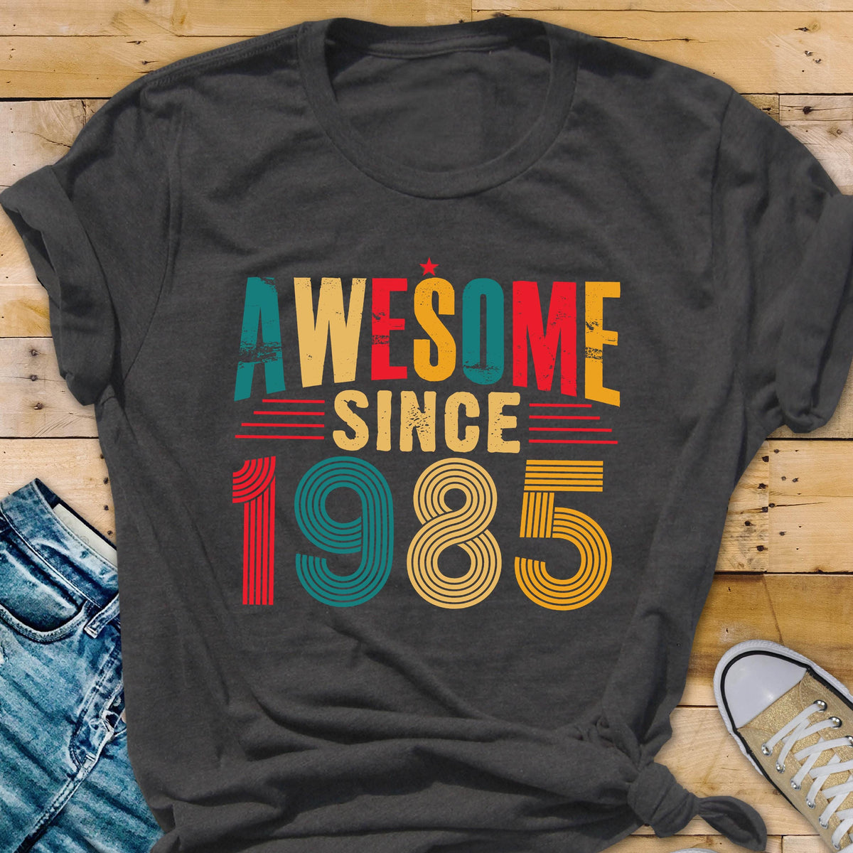 May include: A dark gray t-shirt with the text "AWESOME SINCE 1985" in a retro style with colorful stripes.