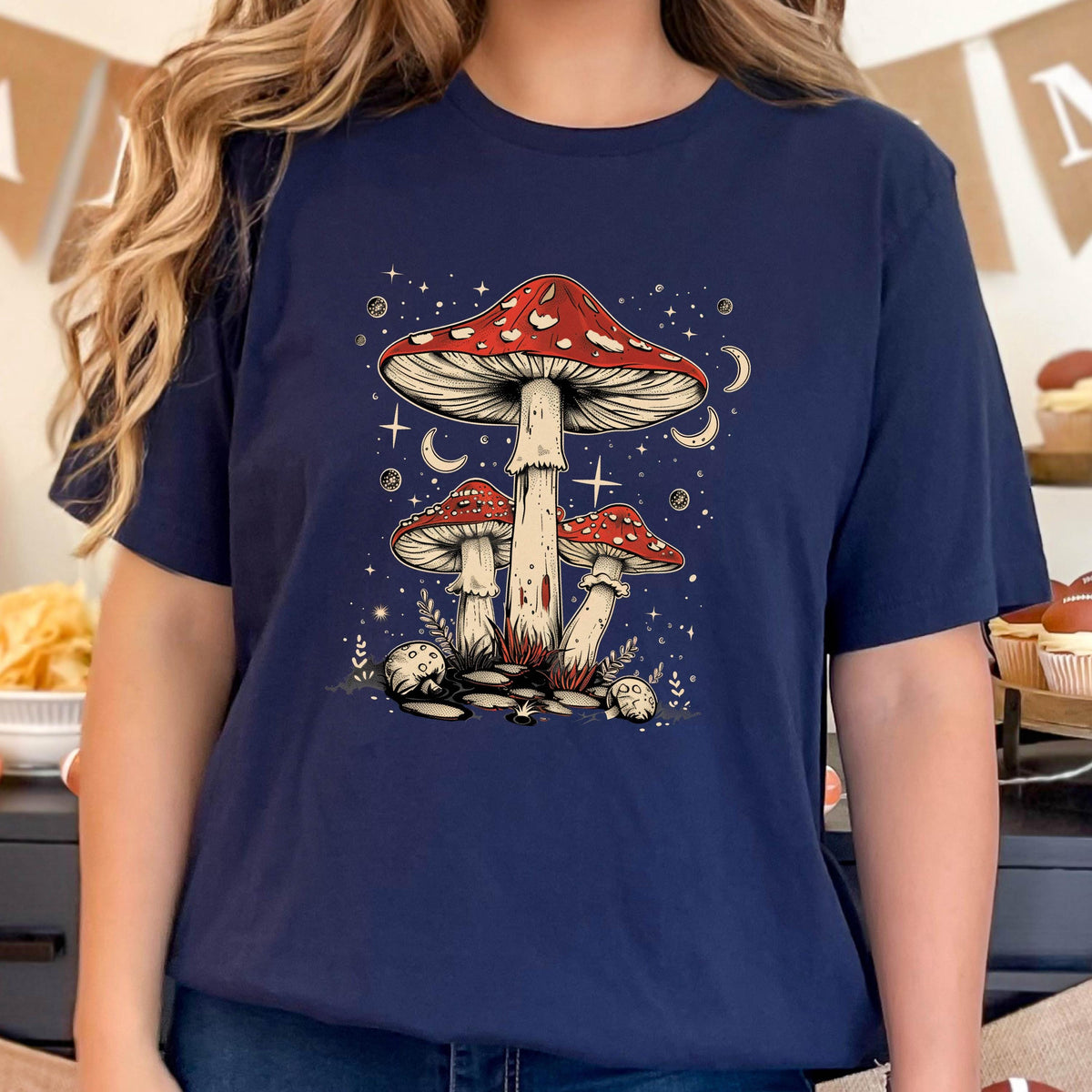 May include: A navy blue t-shirt with a graphic of three red and white mushrooms with white spots. The mushrooms are surrounded by white stars and crescent moons. The text "Shroom Queens" is printed on the shirt.