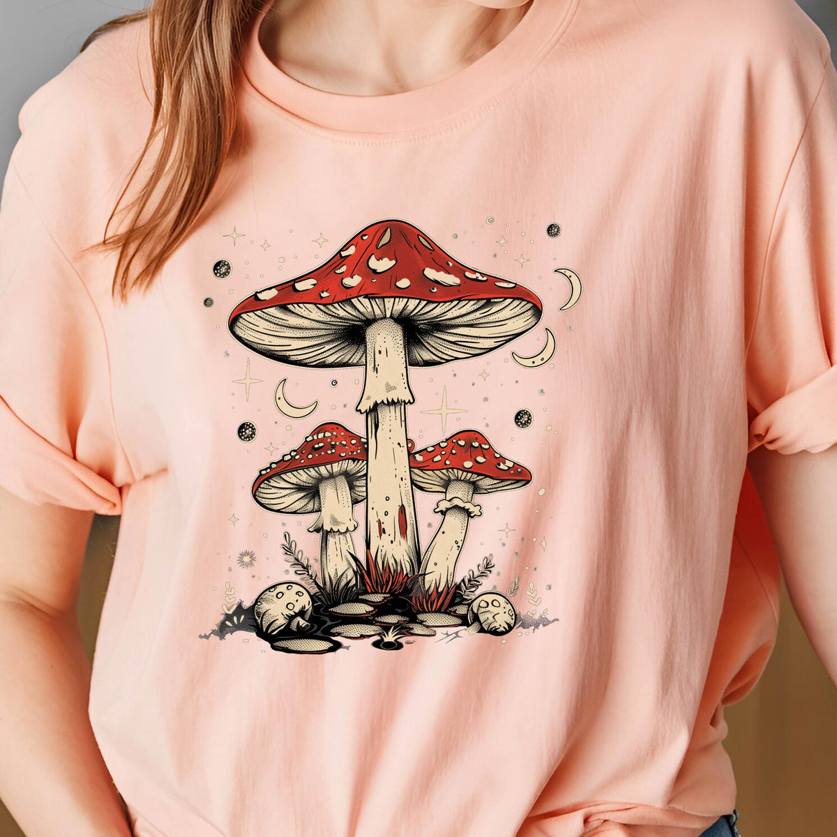 May include: A pink t-shirt with a graphic of three red and white mushrooms with white stems and a crescent moon design. The mushrooms are surrounded by a white starry design.