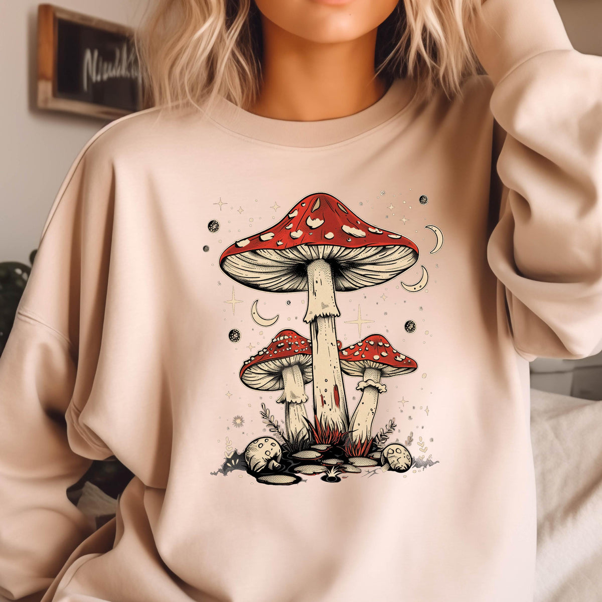 May include: A beige sweatshirt with a graphic print of three red and white mushrooms with white stems. The mushrooms are surrounded by stars and crescent moons.