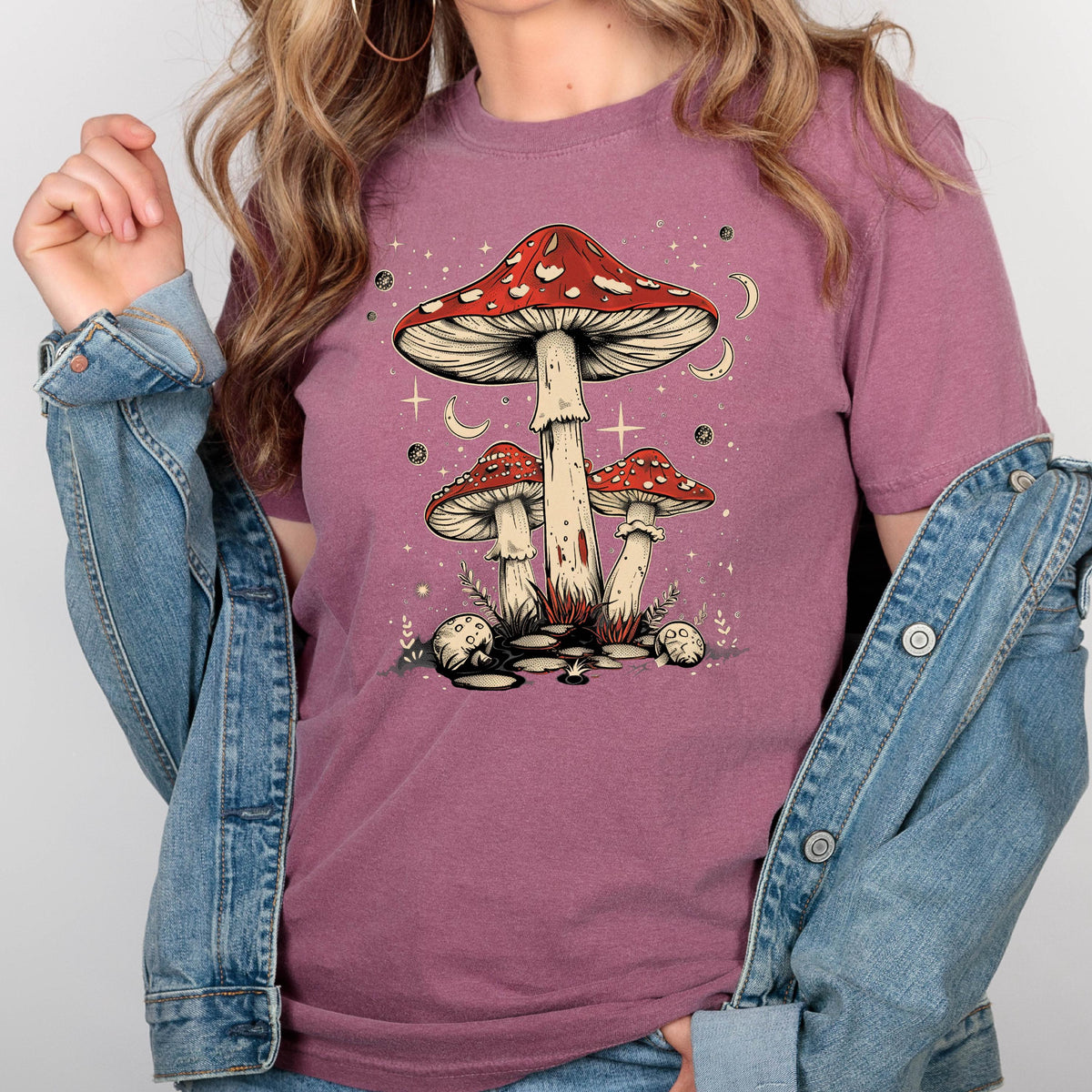 May include: A mauve t-shirt with a graphic of three red and white mushrooms with white spots. The mushrooms are surrounded by white stars and crescent moons. 
