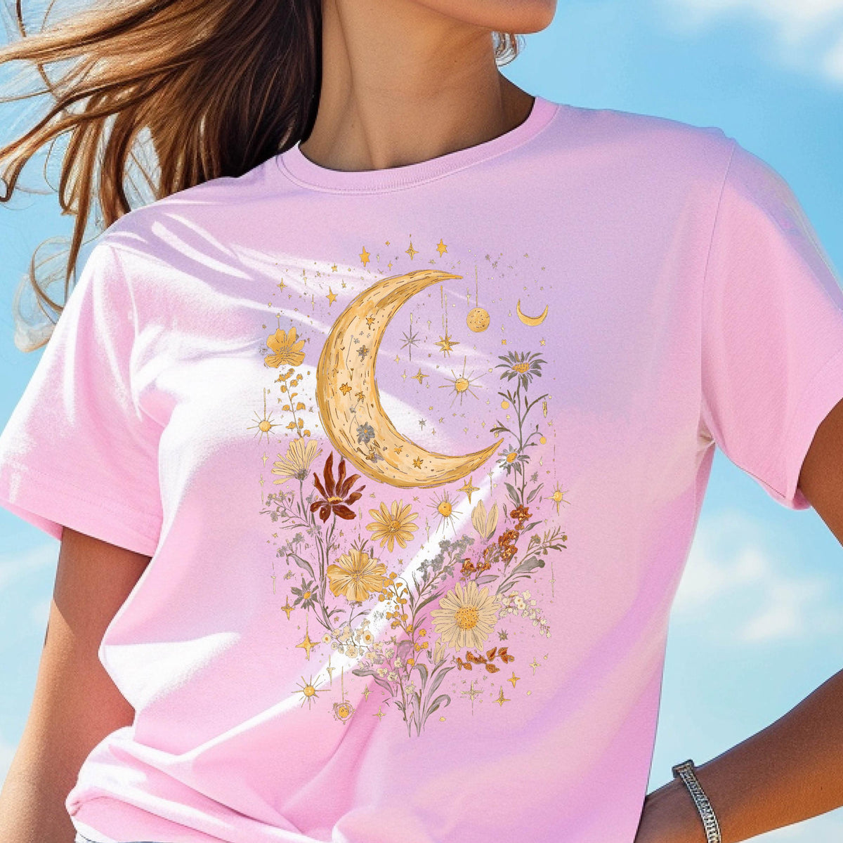 May include: A light pink t-shirt featuring a celestial design. The graphic includes a crescent moon, stars, and various flowers in shades of gold and yellow. The shirt is a crew neck style.