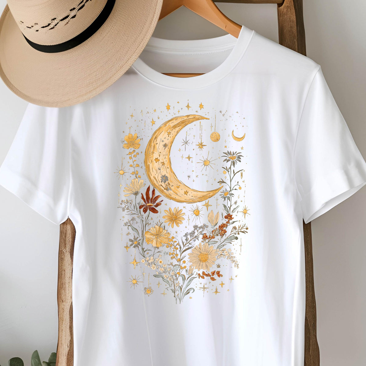 May include: White t-shirt featuring a golden crescent moon design surrounded by yellow and brown flowers, stars, and celestial accents. A tan hat hangs above the shirt. The design has a bohemian or celestial theme.