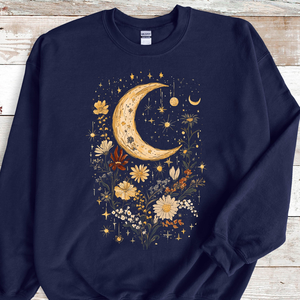 May include: A navy blue crewneck sweatshirt featuring a central graphic print of a large, textured crescent moon surrounded by various wildflowers in shades of yellow, orange, and white, accented with small stars and celestial sparkles throughout.