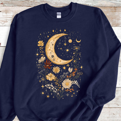 May include: A navy blue crewneck sweatshirt featuring a central graphic print of a large, textured crescent moon surrounded by various wildflowers in shades of yellow, orange, and white, accented with small stars and celestial sparkles throughout.