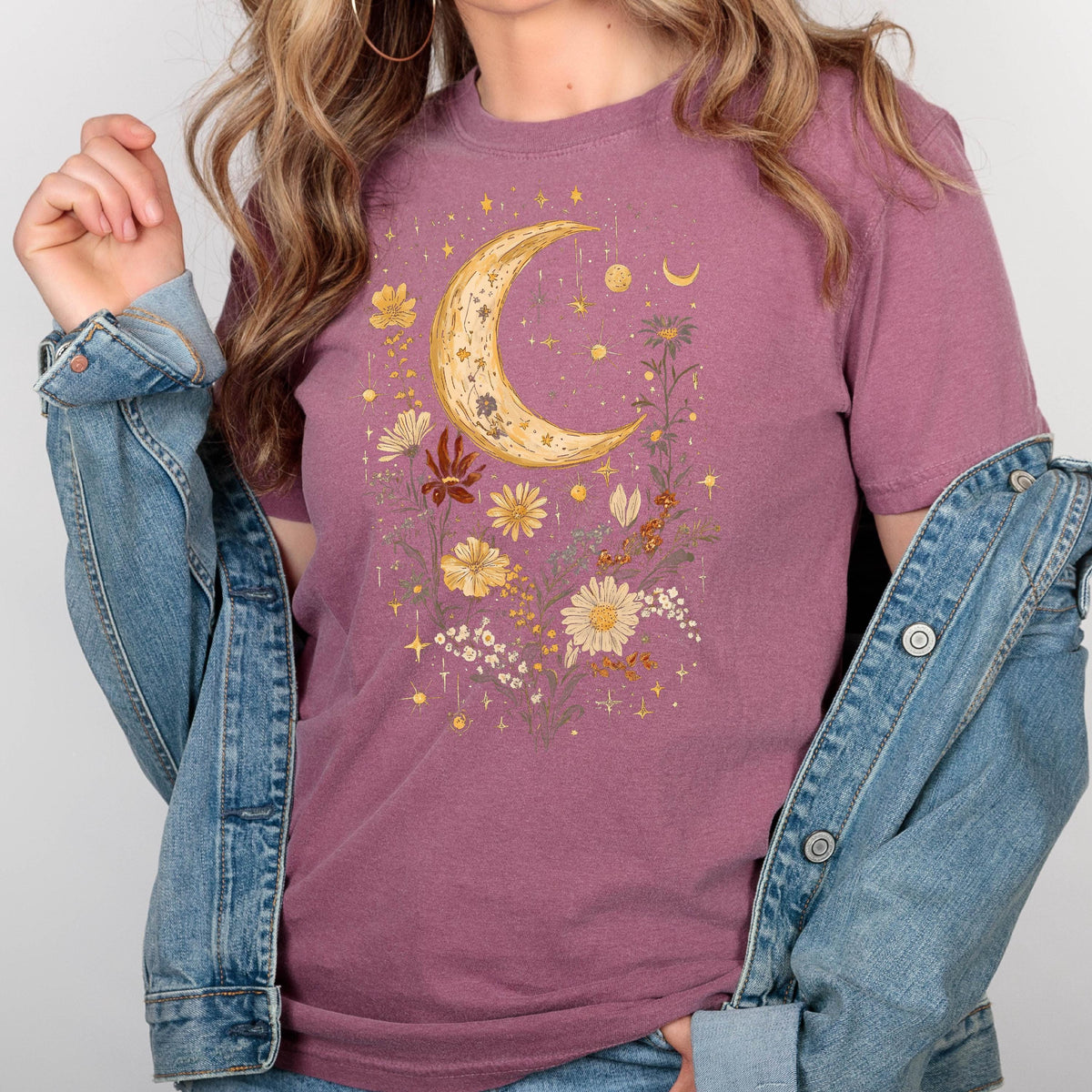 May include: A mauve t-shirt with a crescent moon and floral design in gold and yellow. The design features a crescent moon surrounded by flowers and stars.