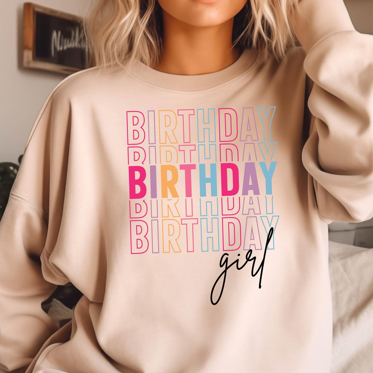 May include: A beige sweatshirt with the text "Birthday girl" in rainbow colors. The text is stacked and repeated multiple times.