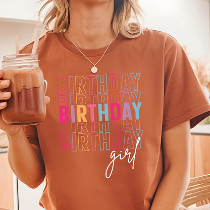 May include: A brown t-shirt with the text "Birthday girl" in rainbow colors. The text is stacked and repeated multiple times.