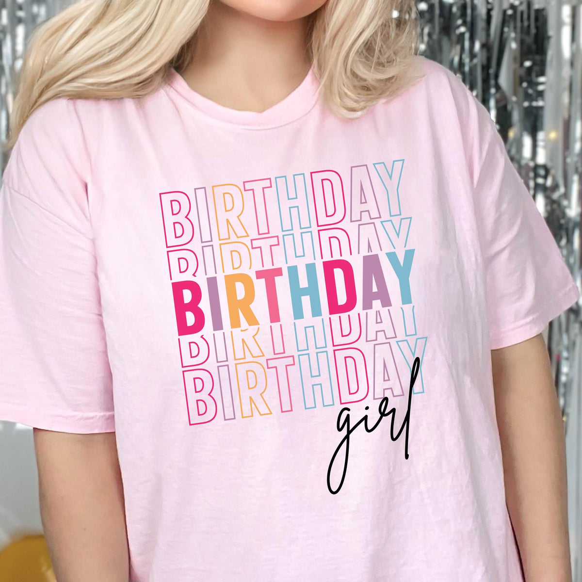 May include: Pink t-shirt with the text "Birthday" repeated in a rainbow color pattern and the text "girl" in black script.