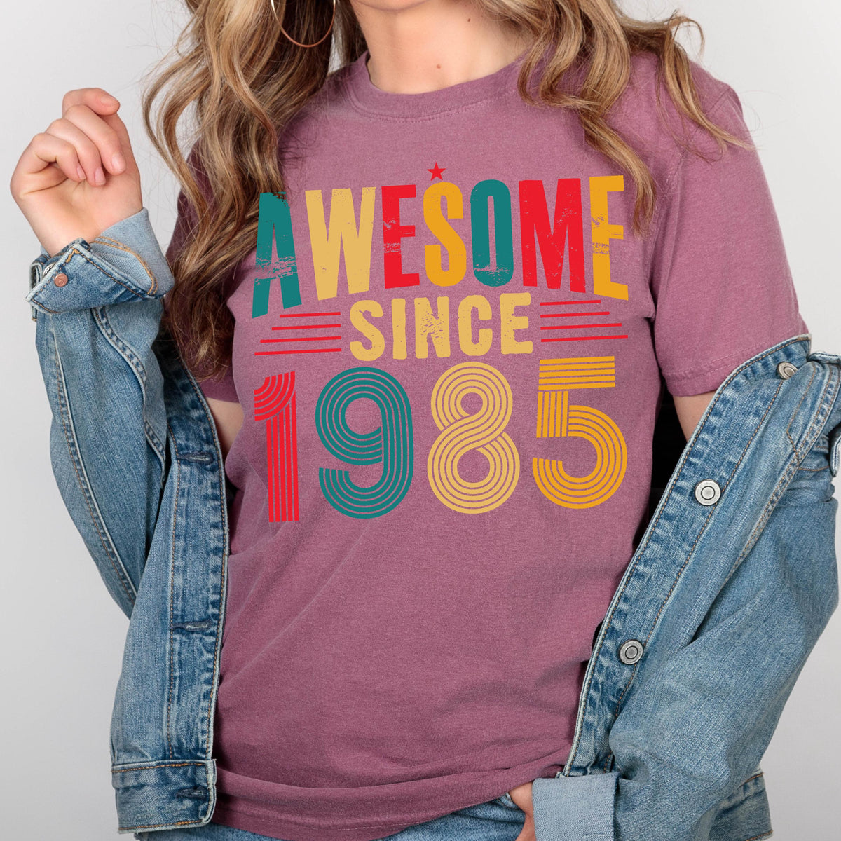 May include: A mauve t-shirt with the text "AWESOME SINCE 1985" in a retro style font with colorful stripes. The numbers are in a rainbow color scheme.
