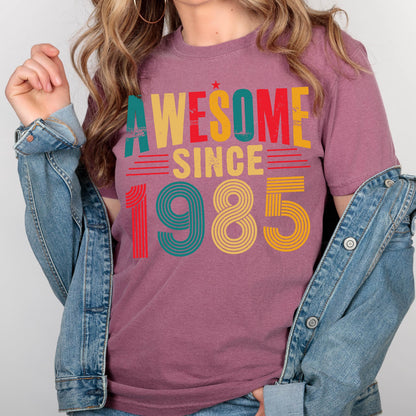 May include: A mauve t-shirt with the text "AWESOME SINCE 1985" in a retro style font with colorful stripes. The numbers are in a rainbow color scheme.