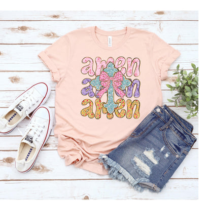 May include: A pink t-shirt with the word "amen" written three times in a colorful, stylized font. A floral cross with a pink bow is in the center of the design.