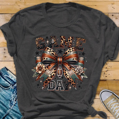 May include: A charcoal gray t-shirt with a graphic print of a leopard print bow with green and orange stripes. The bow is tied around a football. The text "GAME DAY" is printed below the bow.